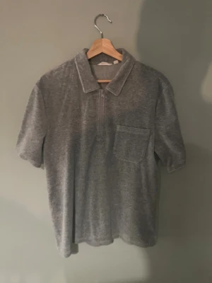 Grå kortärmad polotopp med ficka - Säljer en grå polotopp i mjukt och lent material  med korta ärmar och en bröstficka. Toppen har klassisk krage och en half zip-dragkedja framtill som ger en sportig vibe. Perfekt till jeans eller shorts för en chill look.