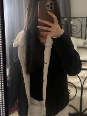 Svart teddyjacka med vit fuskpäls - Säljer en svart teddyjacka med vit, fluffig fuskpäls på insidan och kragen. Jackan har knappar framtill och en klassisk rak passform. Perfekt för dig som vill ha en mysig och stilren look under kyliga dagar.