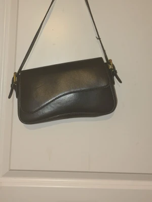 Svart axelväska  - Säljer en stilren svart axelväska i slätt skinn med justerbart axelband och guldfärgade metalldetaljer. Väskan har en minimalistisk design med mjuka former och passar perfekt till dig som gillar en clean look.
