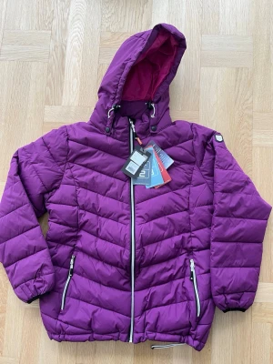 Lila pufferjacka från Tuxer med luva - Säljer en pufferjacka från Tuxer i färgen dark fuchsia med huva och dragkedja framtill. Jackan har två fickor med dragkedja, justerbar dragsko nertill och en snygg rosa insida. Perfekt för kalla dagar och riktigt skön att bära. Jackan klarar 2000 vattenpelare och har hög värmeisolerings förmåga. Jackan är helt ny o kapparna sitter kvar. Nypris 1.099 kr. 