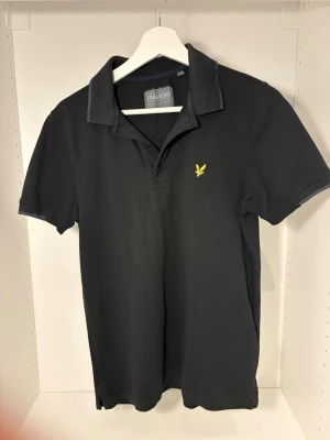 Svart piké från Lyle & Scott - Svart pikétröja från Lyle & Scott med klassisk krage och korta ärmar. Tröjan har gul broderad logga på bröstet och knappar vid halsen. Materialet är mjuk bomull som känns skönt mot huden. Perfekt för en clean och stilren look.