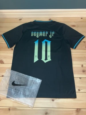 Nike Brasil Neymar Jr. fotbollströja M - Säljer en svart Nike fotbollströja med Neymar Jr. och nummer 10 på ryggen. Tröjan har turkosa detaljer, Brasilien-emblem och Dri-FIT-material som andas. Perfekt för dig som älskar fotboll och vill ha en snygg och bekväm matchtröja.