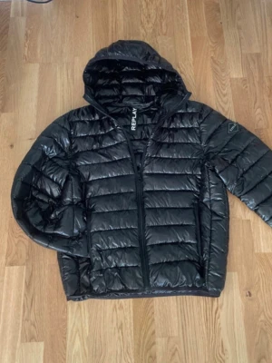 Svart pufferjacka från Replay - Säljer en svart pufferjacka från Replay med huva och dragkedja. Jackan har en glansig finish, quiltad design och Replay-logga på ärmen. Perfekt för kyliga dagar och riktigt snygg till streetwear-stilen.