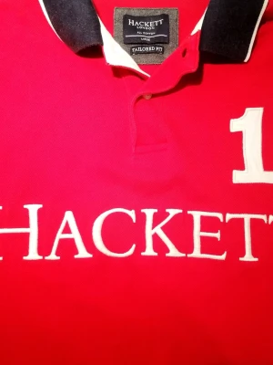 Röd Hackett London piké med brodyr  - Snygg röd pikétröja från Hackett London med stor vit broderad logga och siffran 1 på bröstet. Marinblå krage med vit kant och en vit-en marinblå ärm ger en sportig look. Tillverkad i bomull och har klassisk knappslå framtill.