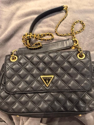 Väska från Guess - Snygg svart axelväska från Guess. Har en kedjerem med inslag av skinn och ett klassiskt Guess-emblem framtill. Pris kan diskuteras 
