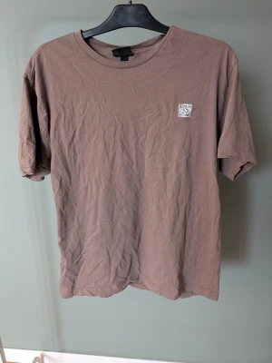 Brun t-shirt med tryck - Säljer en brun t-shirt i mjuk bomull med rund hals och korta ärmar. På bröstet finns en liten QR-kod och på ryggen ett större tryck som liknar en fraktetikett. Perfekt för dig som gillar streetwear med unika detaljer.