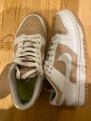 Nike Dunk sneakers beige och vita - Säljer ett par Nike Dunk sneakers i beige och vitt skinn. Klassisk låg modell med vit sula, perforerad tåbox och vita snören. Swoosh-logga på sidan och bekväm vadderad insida. Perfekta för dig som gillar stilrena och neutrala färger.