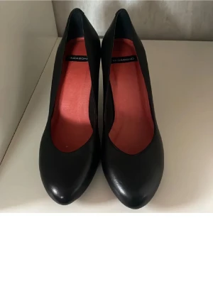 Svarta pumps från Vagabond i skinn - Stilrena svarta pumps från Vagabonder med rund tå och hög klack. Skorna är tillverkade i slätt skinn och har en snygg röd innersula som ger en cool kontrast. Perfekta för dig som vill ha en klassisk men modern look.