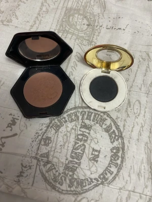 Bronzer och svart ögonskugga duo - Säljer ett set med en matt brun bronzer i sexkantig svart dosa med spegel och en svart ögonskugga i rund vit dosa med guldfärgat lock. Perfekt för att skapa både markerade och sotade looks. Båda produkterna är i puderform.