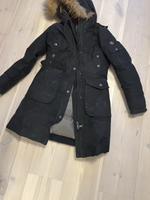 Svart parkas från Canadiens - Säljer en svart parkas från Canadiens med pälsdetalj på huvan och flera stora fickor. Jackan har bälte i midjan, dragkedja och knappar framtill samt broderad logga på insidan. Perfekt för kalla vinterdagar. Nypris 12 tusen. Pris kan diskuteras vid snabb affär