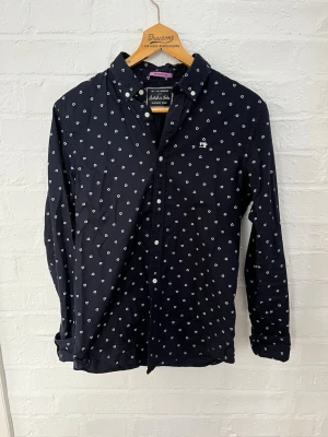Mörkblå prickig skjorta Scotch & Soda - Mörkblå Oxford-skjorta från Scotch & Soda med vita prickar i form av små trianglar. Klassisk krage med knappar och broderad logga på bröstet. Skjortan har långa ärmar och vita knappar framtill. Perfekt för dig som gillar stilrena detaljer.