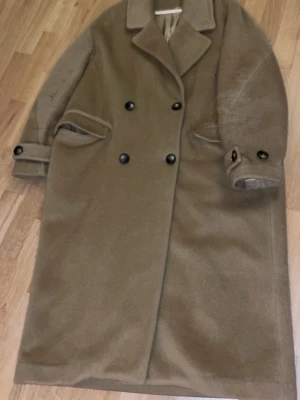 Brun ullkappa med dubbelknäppning - Säljer en klassisk brun kappa i ull med dubbelknäppning och bred krage. Jackan har långa ärmar med knappar vid ärmsluten och två fickor framtill. Perfekt för dig som vill ha en stilren och tidlös look under kyliga dagar. Oversized 