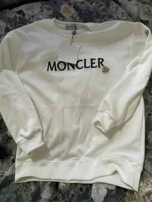 Vit sweatshirt från Moncler - Säljer en clean vit sweatshirt från Moncler med svart logotyptryck på bröstet och klassisk patch på sidan. Tröjan har rund hals, långa ärmar och ribbade muddar. Perfekt för dig som gillar stilrena plagg med exklusiv känsla.