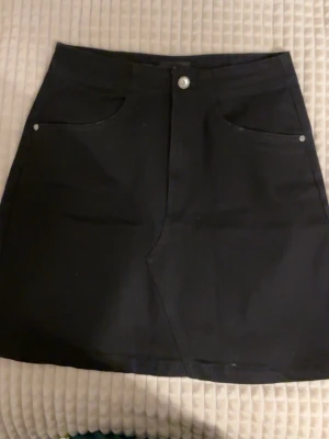 Svart kortkjol med fickor - Säljer en svart kortkjol i klassisk jeansmodell med två framfickor och två bakfickor. Kjolen har knapp och dragkedja framtill och är lätt A-linjeformad. Perfekt basplagg till garderoben.