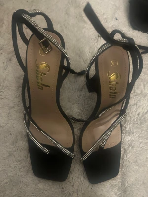 Svarta sandaler med strass från Shein - Snygga svarta sandaler från Shein med fyrkantig tå och tunna remmar dekorerade med glittrande strass. Skorna har klack och knyts runt vristen för en trendig look. Perfekta för att lyfta vilken outfit som helst.