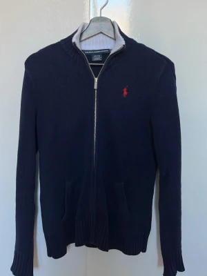 Marinblå stickad kofta från Ralph Lauren - Säljer en marinblå stickad kofta från Ralph Lauren med dragkedja framtill och ribbade muddar. Klassisk röd logga på bröstet och två fickor fram. Perfekt till höst och vinter för en clean och stilren look.