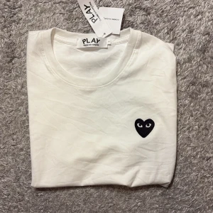 Vit t-shirt från Comme des Garçons Play - Säljer en vit t-shirt från Comme des Garçons Play med det ikoniska svarta hjärtat med ögon broderat på bröstet. T-shirten har rund hals och är kortärmad, tillverkad i mjuk bomull. Perfekt för dig som gillar stilrena och coola detaljer.