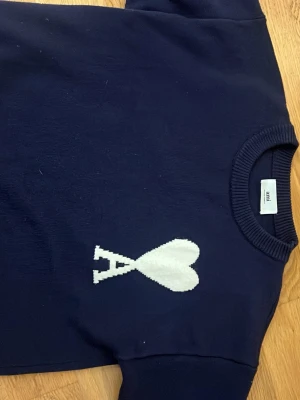 Mörkblå stickad tröja från Ami - Säljer en mörkblå stickad tröja från Ami Alexandre Mattiussi med vit hjärtlogga och bokstaven A på bröstet. Tröjan har rund halsringning och ribbade muddar. Perfekt för dig som gillar stilrena detaljer och vill ha något som sticker ut. Förlåt för dåliga bilder
