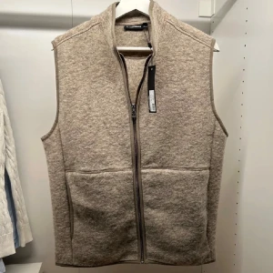 Beige ullväst från J.Lindeberg - Säljer en stilren beige ullväst från J.Lindeberg med hög krage och hel dragkedja framtill. Västen har två fickor och en diskret logga i nacken. Perfekt att slänga över en hoodie eller skjorta för extra värme.