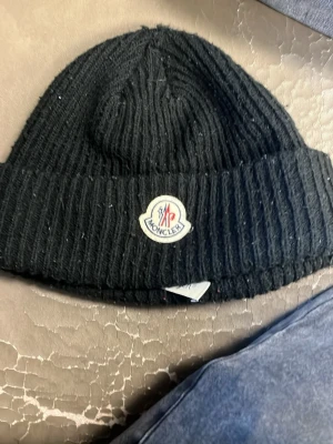 Svart Moncler mössa i ull - Ribbstickad svart mössa från Moncler med klassisk logotyp framtill. Tillverkad i mjuk ull som håller dig varm under kyliga dagar. Enkel och stilren design som passar till det mesta. Priset går diskutera 