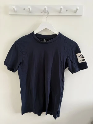 Herr Mörkblå Umbro t-shirt med logga - Säljer en mörkblå t-shirt från Umbro i storlek S. Klassisk passform med korta ärmar och rund hals. Stor vit Umbro-logga tryckt på ena ärmen ger en sportig vibe. Är skön och ha på sig. 