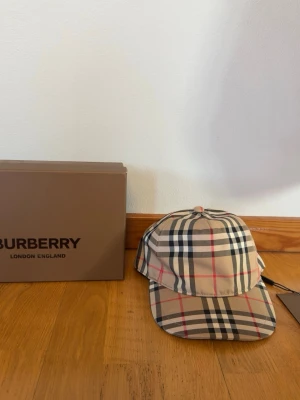 Rutig Burberry keps i beige och svart - Säljer en klassisk Burberry keps i storlek M med det ikoniska rutiga mönstret i beige, svart, vitt och röda detaljer. Kepsen har justerbar baksida och Burberry-logga bak. Materialet är en mix av bomull och polyester, perfekt för dig som vill ha en stilren och tidlös accessoar.