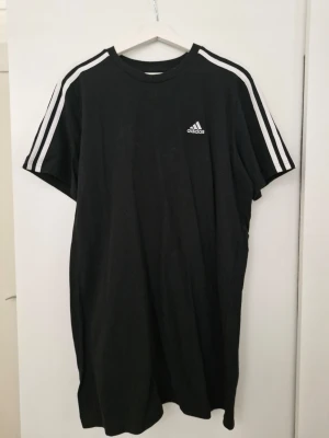 Svart Adidas t-shirtklänning L - Svart t-shirtklänning från Adidas med klassiska vita ränder på axlarna och Adidas-logga på bröstet. Modellen är loose fit och har korta ärmar. Perfekt för en sportig och avslappnad stil.