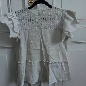 Vit romantisk topp - Vit topp i lätt, krinklat tyg med vackra broderade detaljer framtill och volangärmar. Knäppning baktill i nacken. Toppen är i fint använt skick med normal användningskänsla/veck från förvaring. 