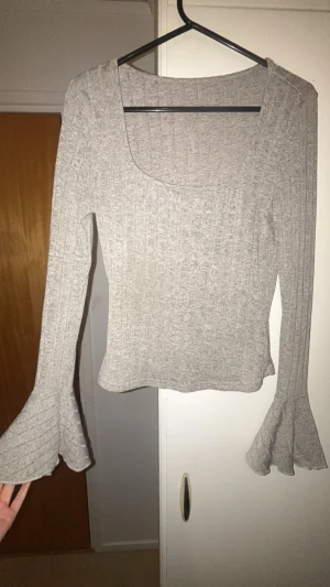 Grå ribbad topp - Trendig grå ribbad topp med fyrkantig ringning och långa trumpetärmar. Croppad modell som ger en snygg siluett. Ett litet hål i tröjan men inget som syns eller märks mycket när den sitter på.