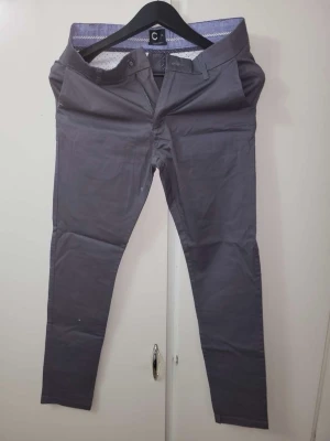Mörkgrå chinos från C - Snygga mörkgrå chinos från C med slim fit passform och klassisk design. Byxorna har två sidofickor, bälteshällor och knappgylf. Tillverkade i ett mjukt och stretchigt bomullsmaterial som ger en skön känsla hela dagen.