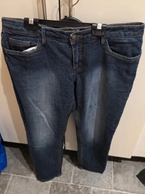 Mörkblå raka jeans med slitningar - Säljer ett par mörkblå jeans med raka ben och snygga slitningar framtill. Jeansen har klassisk femficksdesign och kontrastsömmar på bakfickorna. Materialet är denim i bomull. Perfekta för en avslappnad och trendig look.