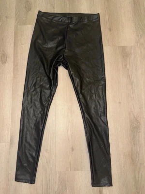 Svarta fuskskinnleggings från H&M - Snygga svarta leggings i fuskskinn från H&M. De har hög midja och smal passform som sitter tight längs benen. Materialet är glansigt och ger en cool look. Perfekta att matcha med oversized tröjor eller crop tops för en trendig outfit.