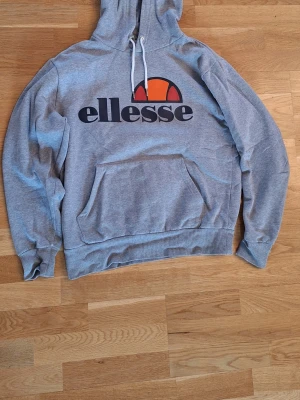 Grå Ellesse hoodie med logga - Snygg grå hoodie från Ellesse med stor svart logga och orange detalj på bröstet. Klassisk känguruficka framtill, vit dragsko i huvan och mjukt material på insidan. 