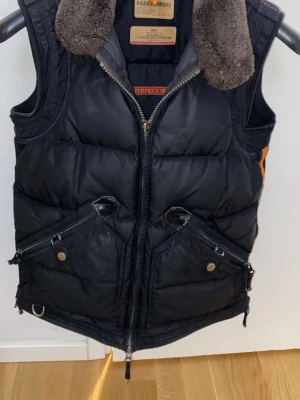 Svart dunväst från Parajumpers - Svart dunväst från Parajumpers med två stora snedställda fickor med dragkedja och tryckknapp, samt en mjuk brun fuskpälskrage. Västen har markerade sömmar och orange detalj på sidan. Perfekt för lager-på-lager-stil.