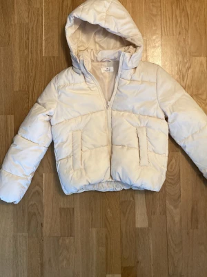 Vit pufferjacka med huva från H&M - Snygg vit pufferjacka från H&M med huva och dragkedja framtill. Jackan har två stora fickor på sidorna och elastisk nederkant. Perfekt för kalla vinterdagar och riktigt skön att bära. Materialet är syntetiskt och jackan är vadderad för extra värme.