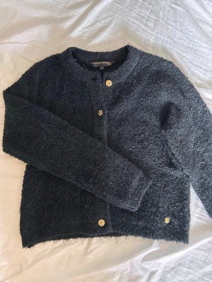 Svart bouclékofta med guldknappar - Mysig svart med viss blå ton bouclékofta från Pernilla Wahlgren med rund halsringning och fem dekorativa guldknappar framtill. Koftan har lång ärm och en fluffig, texturerad yta som ger en trendig look. Perfekt att slänga över en topp för extra stil. Aldrig använt!💗