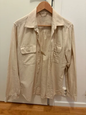 Beige linneskjorta från A Day's March - Säljer en stilren beige skjorta från A Day's March i 100% linne. Skjortan har två bröstfickor med knappar, klassisk krage och långa ärmar. Perfekt för dig som gillar en avslappnad men snygg look.