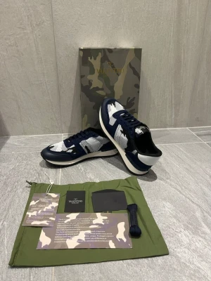 Valentino blåvita sneakers - Snygga sneakers från Valentino i blått och vitt med kamouflagemönster. Skorna har en låg silhuett, mesh- och läderdetaljer samt vita och mörkblå partier. Kommer med originalkartong, dustbag och extra skosnören. Perfekta för dig som gillar exklusiv streetwear.