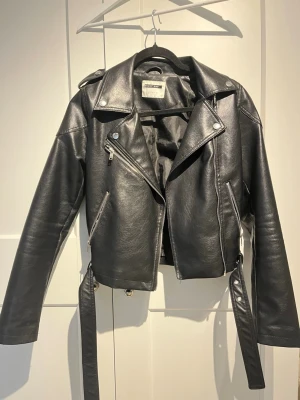 Svart skinnjacka från Bershka - Snygg svart skinnjacka från Bershka med klassisk bikerstil, sned dragkedja, axelklaffar och bälte i midjan. Jackan har silverfärgade detaljer och flera fickor med dragkedja. Perfekt för en cool och trendig look.