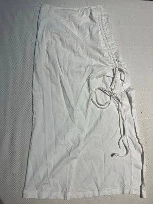 Vit långkjol med snörning i sidan - Trendig vit långkjol med snygg snörning längs sidan som ger en cool detalj. Kjolen har en rak passform och är gjord i ett mjukt, lätt material som känns skönt mot huden. Perfekt för dig som vill ha en stilren men ändå unik look.