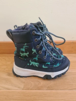 Mörkblå kängor med dinosaurier - Säljer ett par mörkblå kängor med grönt dinosauriemönster och kraftig vit sula från Gulliver i storlek 23. Kängorna har snörning med reflexdetaljer och dragkedja på sidan för enkel påtagning. Insidan är fodrad för extra värme och komfort. Perfekta för kalla dagar! Kängorna är även vattentäta. Endast använda ett fåtal gånger under en säsong så finns inget som helst slitage. 