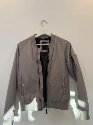 Grå bomberjacka The Black Label - Snygg grå bomberjacka från The Black Label Collection med ribbade muddar och krage. Jackan har två framfickor med tryckknappar och en ärmficka med dragkedja. Tillverkad i ett slätt, lätt material med svart innerfoder och klassisk bomberstil.