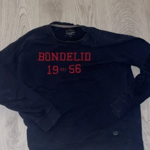 Mörkblå sweatshirt från Bondelid - Mörkblå sweatshirt från Bondelid med röd broderad text 'BONDELID 1956' på bröstet. Tröjan har rund halsringning, ribbade muddar och ett diskret mönster i tyget framtill. Perfekt för en avslappnad och sportig stil. 