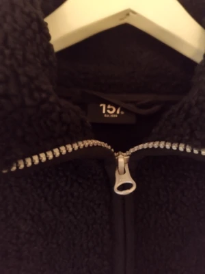 Svart teddyjacka från 157 - Mysig svart teddyjacka från 157 med dragkedja framtill och hög krage. Jackan har en fluffig och mjuk struktur i teddy-material och raka ärmar. Perfekt för kalla dagar när du vill hålla dig varm och snygg.