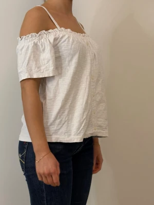 Vit offshoulder topp med knappar - Snygg vit offshoulder topp med korta ärmar och smala axelband. Toppen har knappar framtill och en rynkad kant upptill som ger en avslappnad och trendig look. Perfekt för varmare dagar och enkel att matcha med jeans eller kjol.