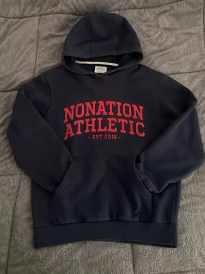 Mörkblå hoodie Nonation Athletic - Mörkblå hoodie med röd text 'NONATION ATHLETIC EST 2015' på bröstet. Klassisk känguruficka framtill och huva. Perfekt för en sportig och avslappnad stil.