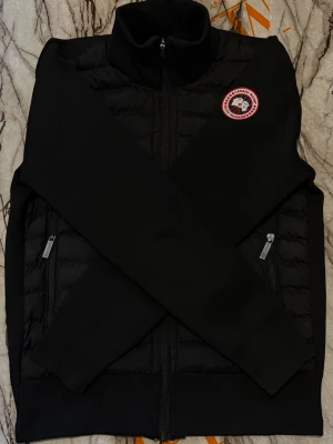 Svart cardiganjacka från Canada Goose - Svart cardiganjacka från Canada Goose med quiltad front, hög krage och dragkedja. Jackan har två fickor med dragkedja och en ikonisk rund logotyp på bröstet. Perfekt för lager-på-lager och snygg streetstil. Kan passa s också 