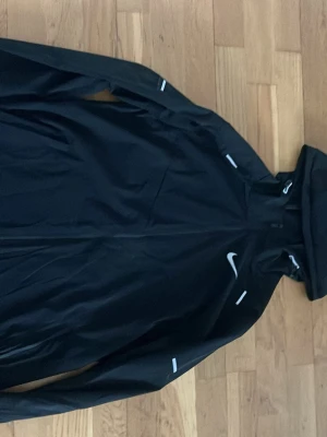 Svart vindjacka från Nike - Snygg svart vindjacka från Nike med vit logga på bröstet och reflekterande detaljer. Jackan har dragkedja framtill och hög krage. Perfekt för blåsiga dagar och sportiga outfits.
