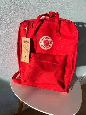 Röd Fjällräven Kånken ryggsäck mini - Säljer en klassisk röd Fjällräven Kånken mini ryggsäck i slitstarkt material. Ryggsäcken har ett stort huvudfack, en framficka med dragkedja och två sidofickor. Ikonisk rund logotyp framtill och praktiska handtag upptill. Helt ny med prislapp.