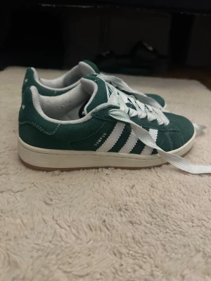 Adidas Campus gröna sneakers mocka - Gröna Adidas Campus sneakers i mocka med klassiska vita ränder på sidorna och vita skosnören. Skorna har en låg profil och vit sula, perfekt för en clean och sportig look. Ikonisk Adidas-logga på plösen och Campus-tryck på sidan.